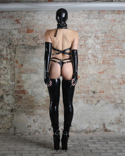 Person wearing a black latex and leather outfit with a hood, harness, and boots against a brick wall.