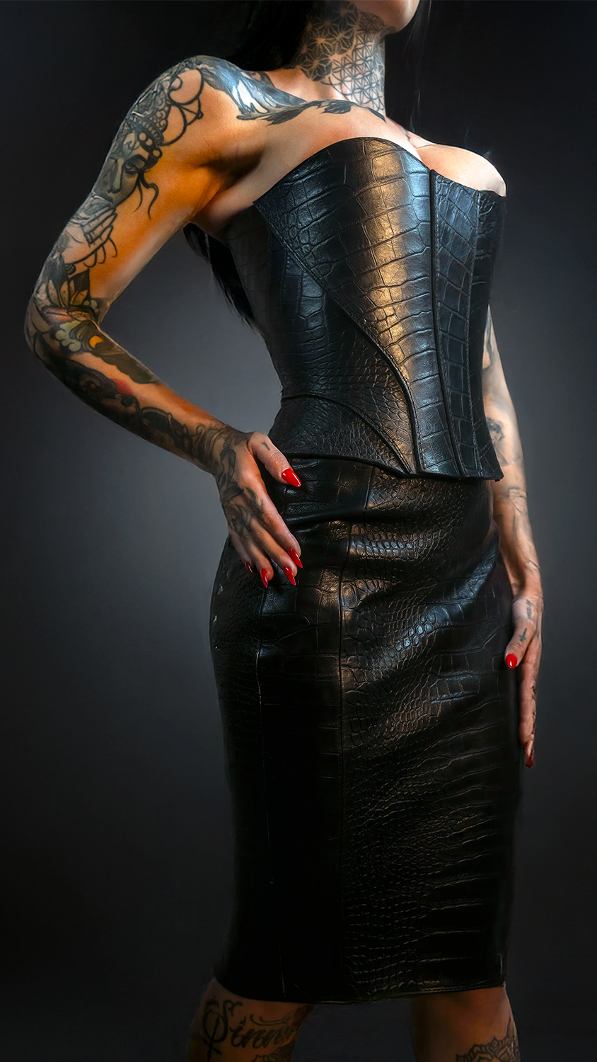 Person wearing a black leather outfit with tattoos on arms and back against a dark background